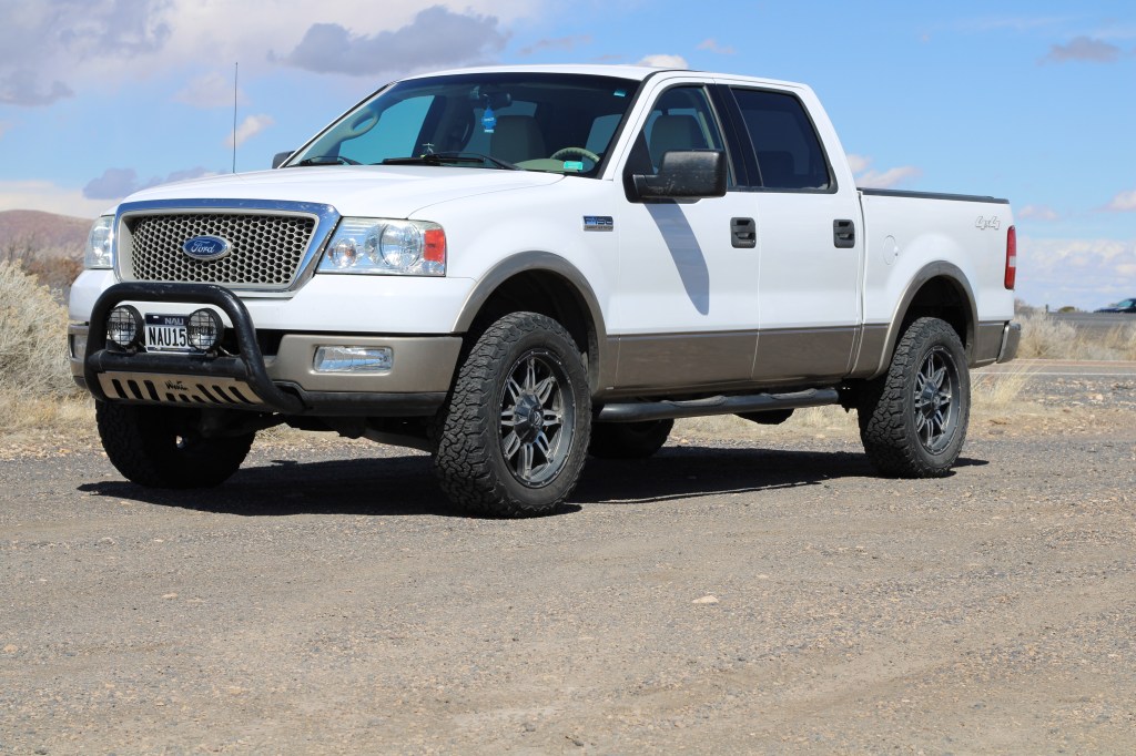 A white Ford pickup truck with custom wheels parked on a gravel road under a blue sky with clouds.