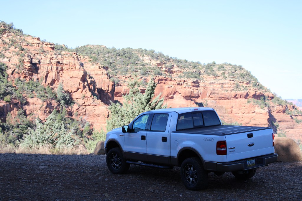 A white pickup truck parked near red rock formations under a clear blue sky.
